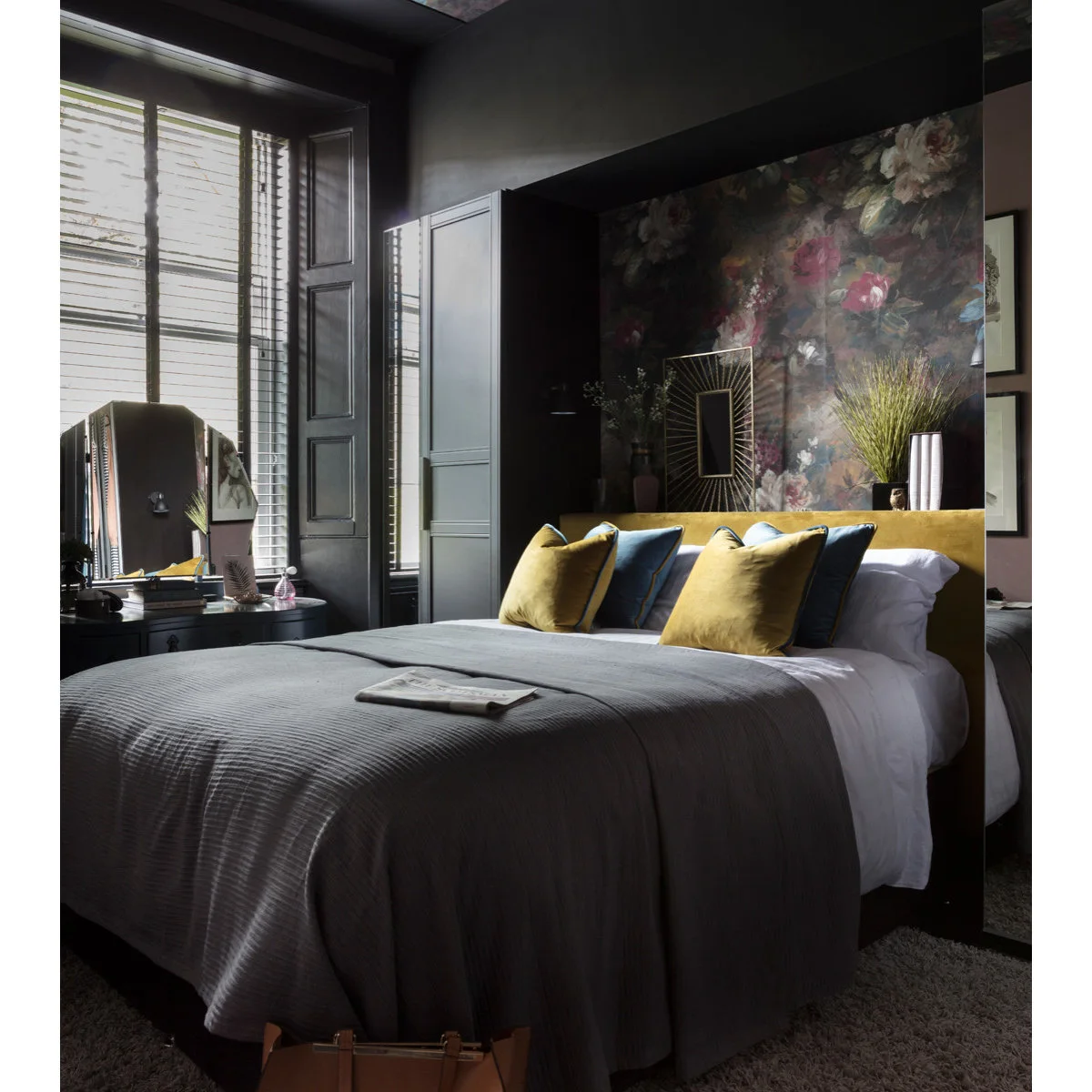 Dark floral wallpaper in a recess above a mustard headboard with narrow mirrors at its sides.