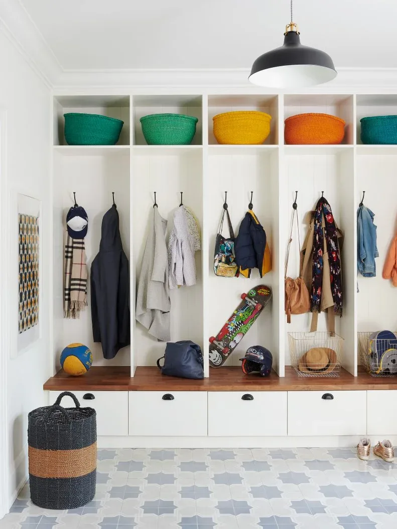 White mudroom cubbies with colorful upper baskets, wooden bench surface, lower drawers, coat hooks and patterned pale floor.