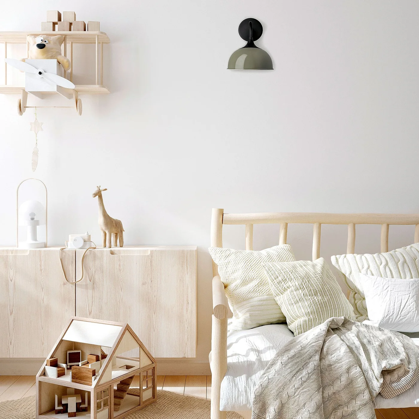 A pale wood bed and matching flat-front cabinet stand against white walls with green and white cushions, a small dollhouse and wall-mounted light
