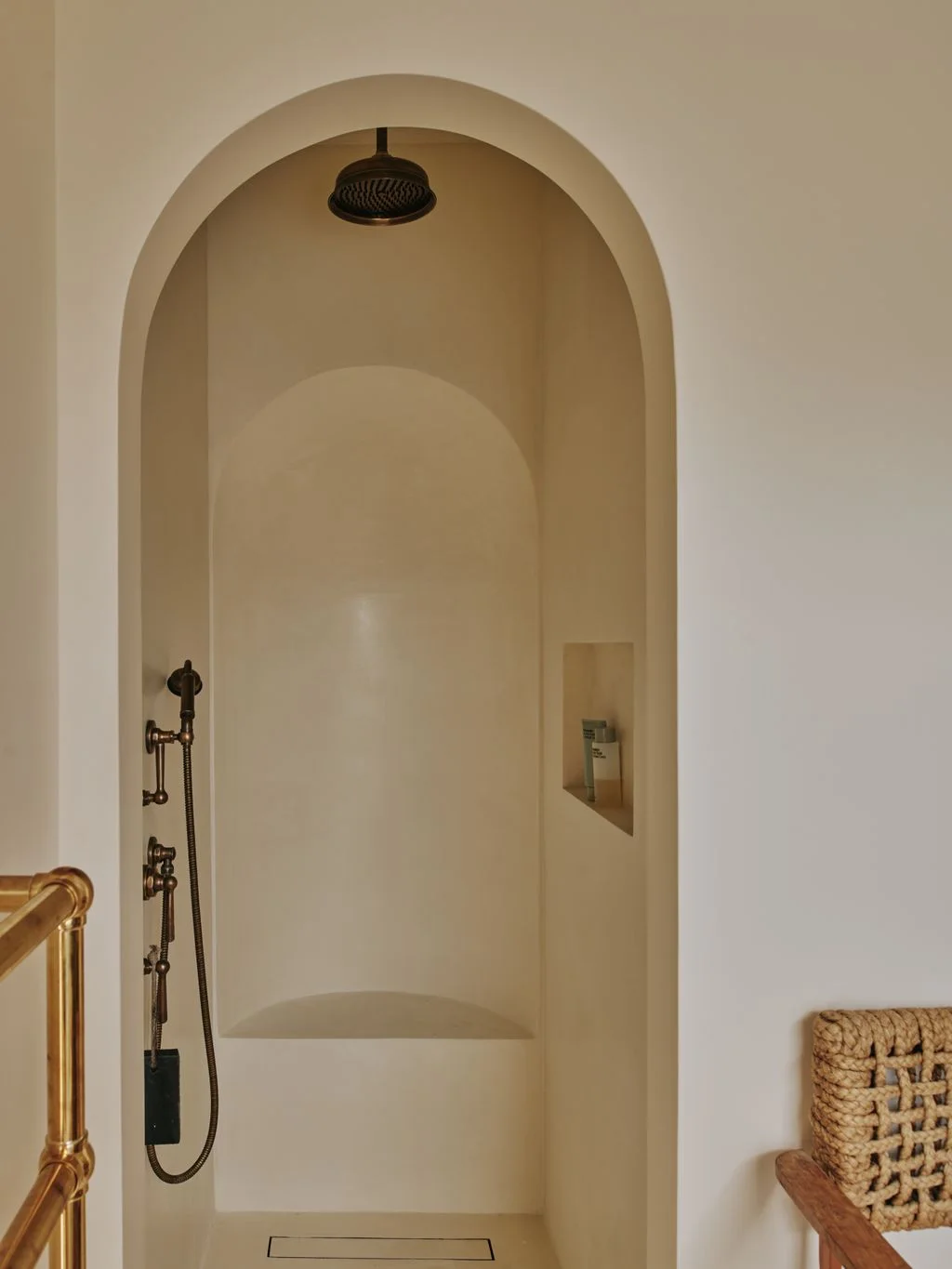 A softly arched open shower has warm pale walls, brass-colored fittings, a recessed shelf and a linear floor drain.