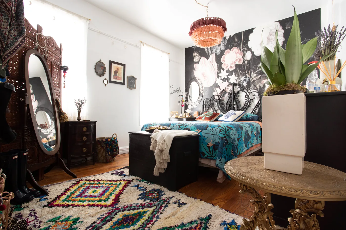 Bedroom with ornate dark headboard, large floral wall mural, turquoise botanical bedding, colorful rug, carved furniture and green potted plant.