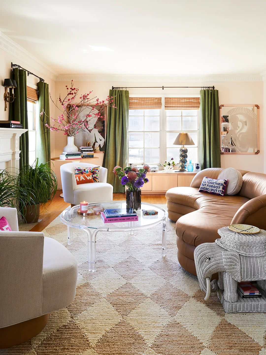Curved tan leather couch in a pale pink room with green curtains, pale chairs and a transparent coffee table.