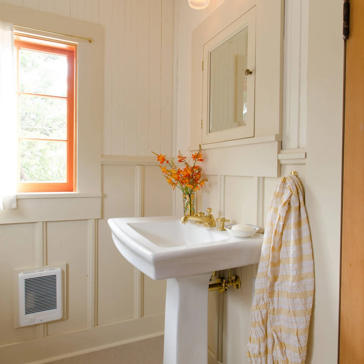 A white pedestal sink sits below a recessed mirror cabinet against pale paneled walls beside a wood-trimmed window.