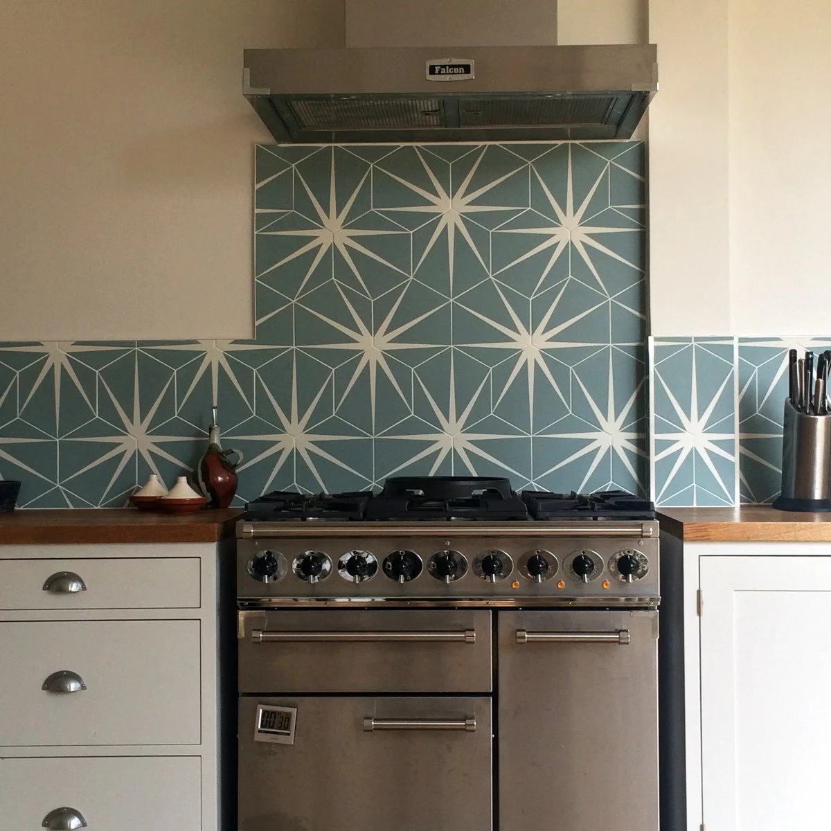 Terrace-house cooking area with blue geometric tiles, pale cabinets and a metal-looking range hood