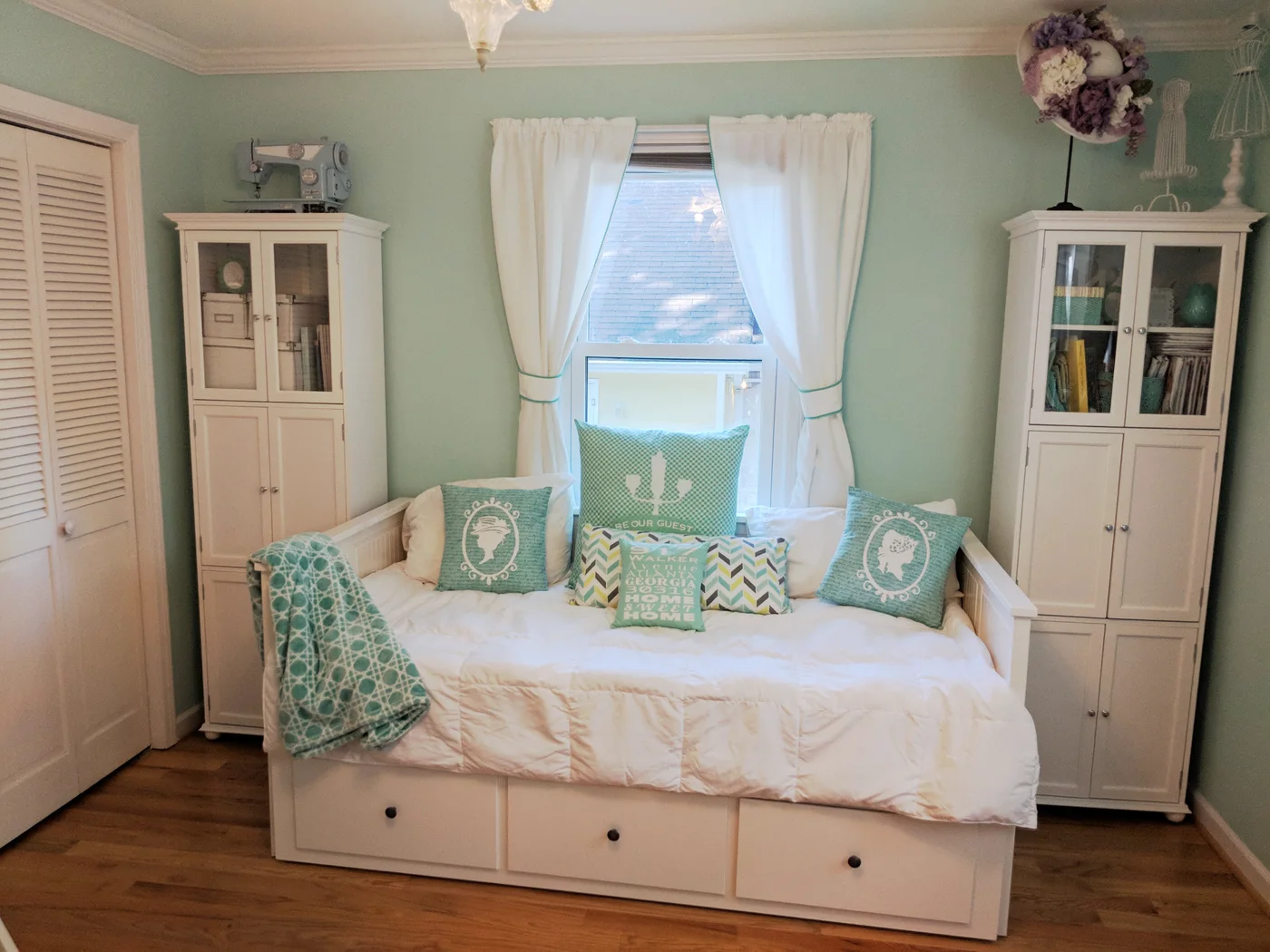 A white daybed with three lower drawers sits between two tall white cabinets in a mint-colored room with patterned cushions.