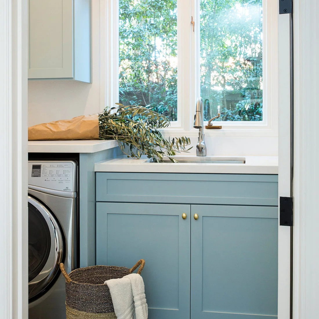 Pale blue cabinets with small warm-metal knobs surround a white utility sink below a bright window beside a laundry machine.