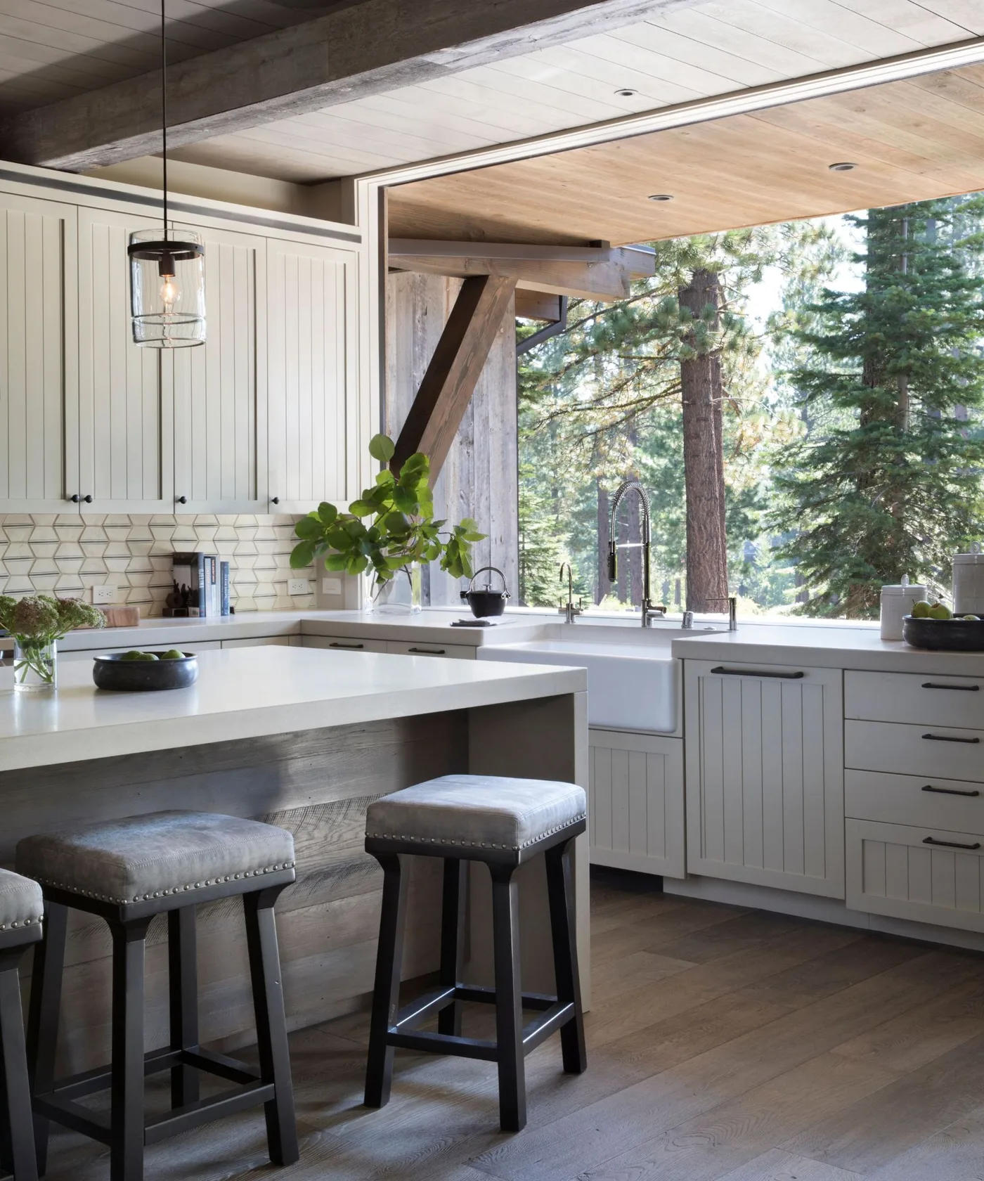 Light kitchen with gray island, wooden stools, white cabinet doors and broad opening toward a windowed room.