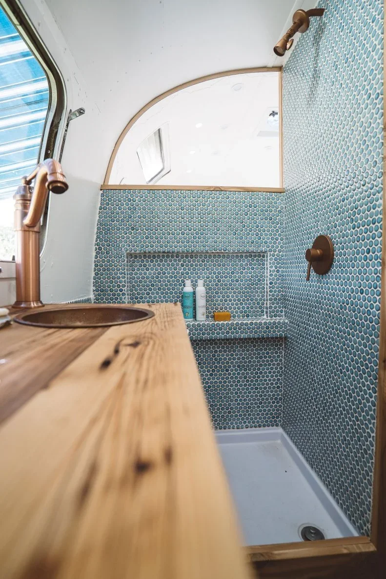 A wood counter and small round basin beside a compact blue penny tile shower