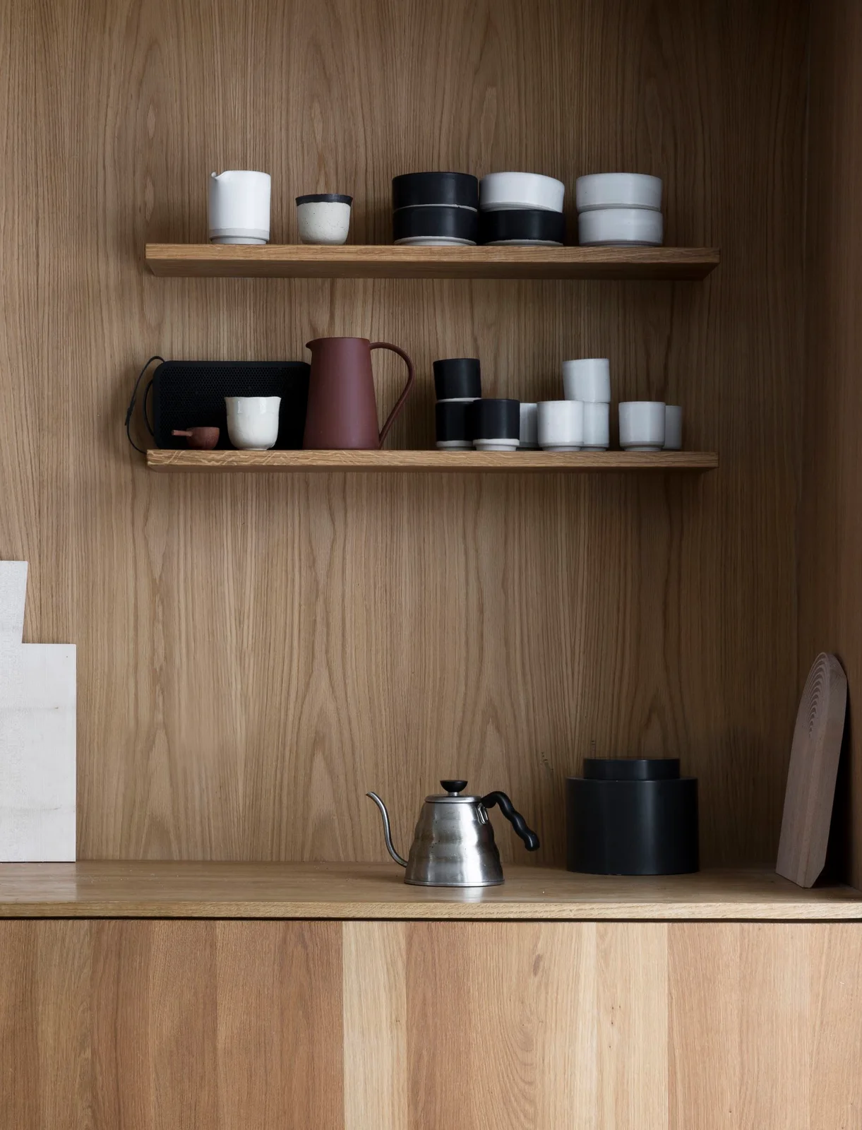 Two wooden shelves hold dark and pale cups above a wooden counter with a metal kettle, dark canisters and cutting boards