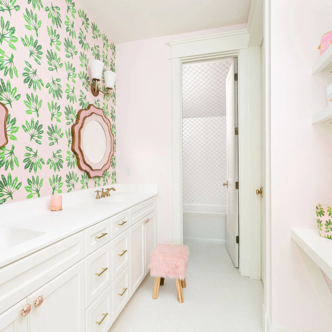 Green leaf wallpaper surrounds shaped pink mirrors above white cabinetry beside pale pink walls and a pink stool.