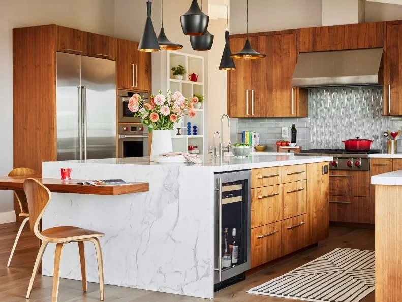 Walnut cabinets surround a white waterfall island with a lower wood dining ledge, metal appliances and mismatched black pendants.