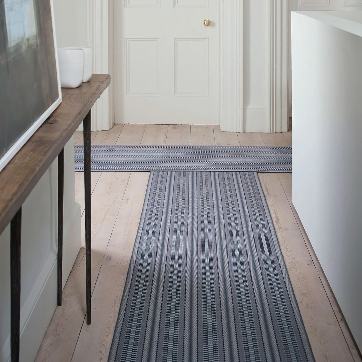 Two pale gray striped runners meeting at a right-angle hallway turn beside a slim wood console and pale doors