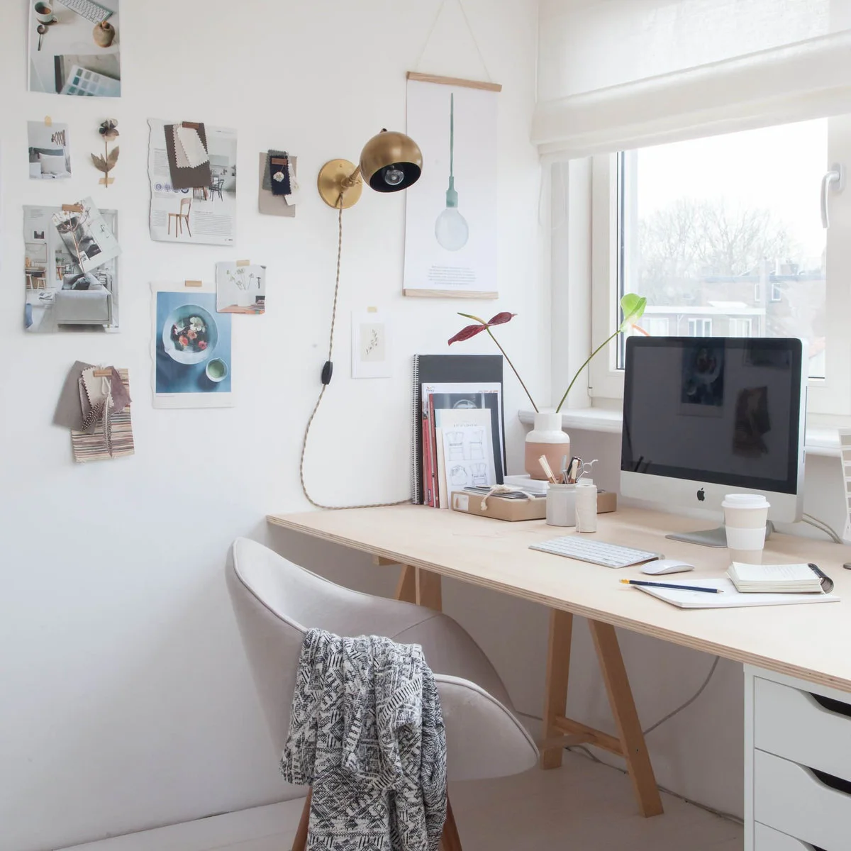 Pale home office with trestle desk, wall-mounted gold-toned lamp, upholstered chair and pinned pictures
