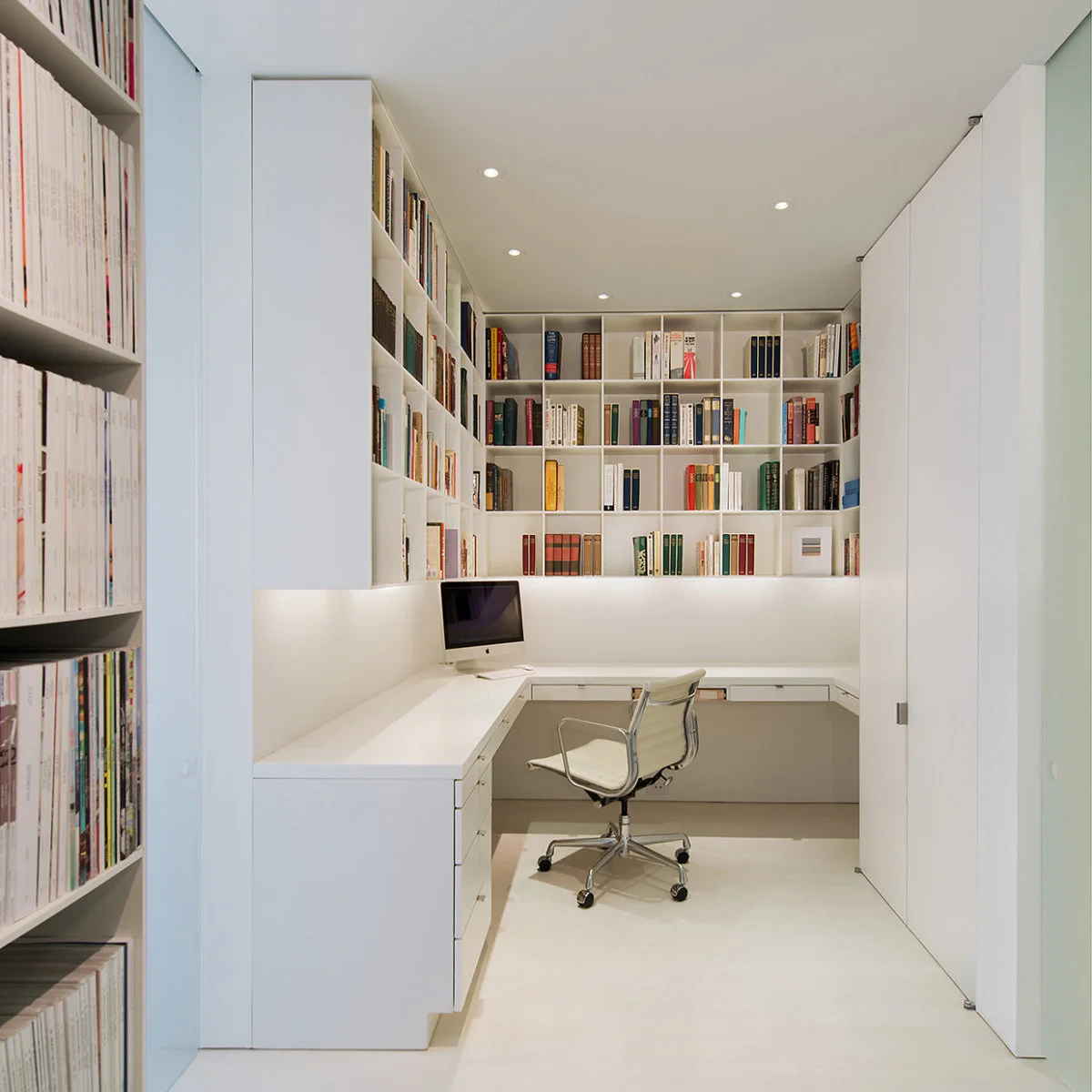 White narrow office nook with bookshelves above an L-shaped desk and closed cupboards to one side