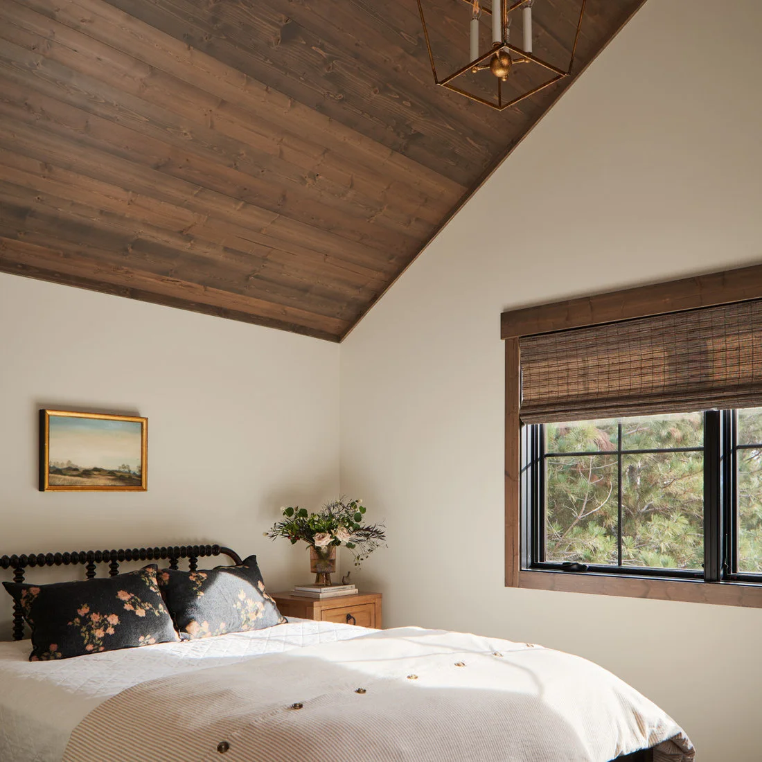 A dark spindle bed with floral cushions below a timber ceiling beside a wooden bedside cabinet