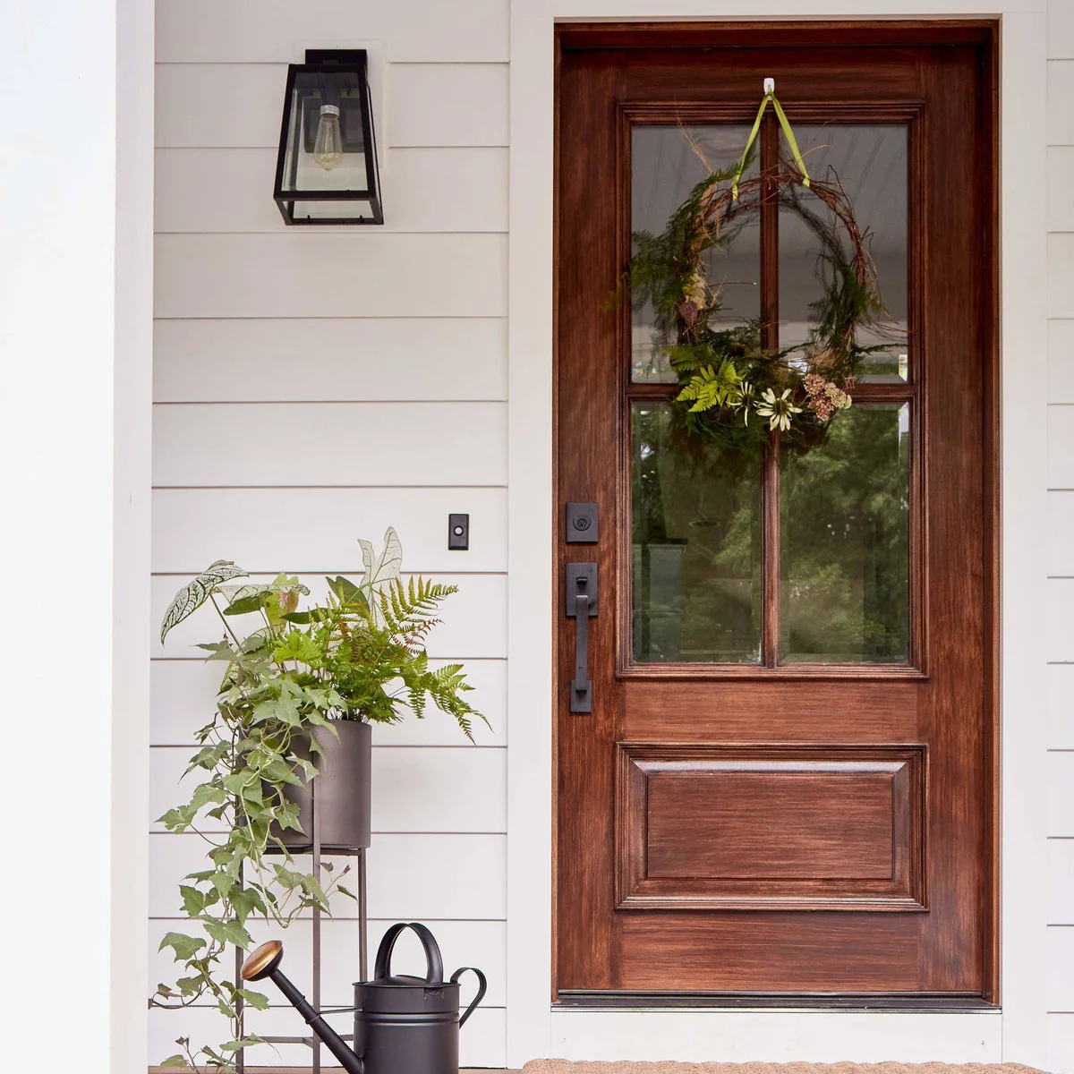 Wood-colored front door beside a raised dark planter, wall lantern and watering can