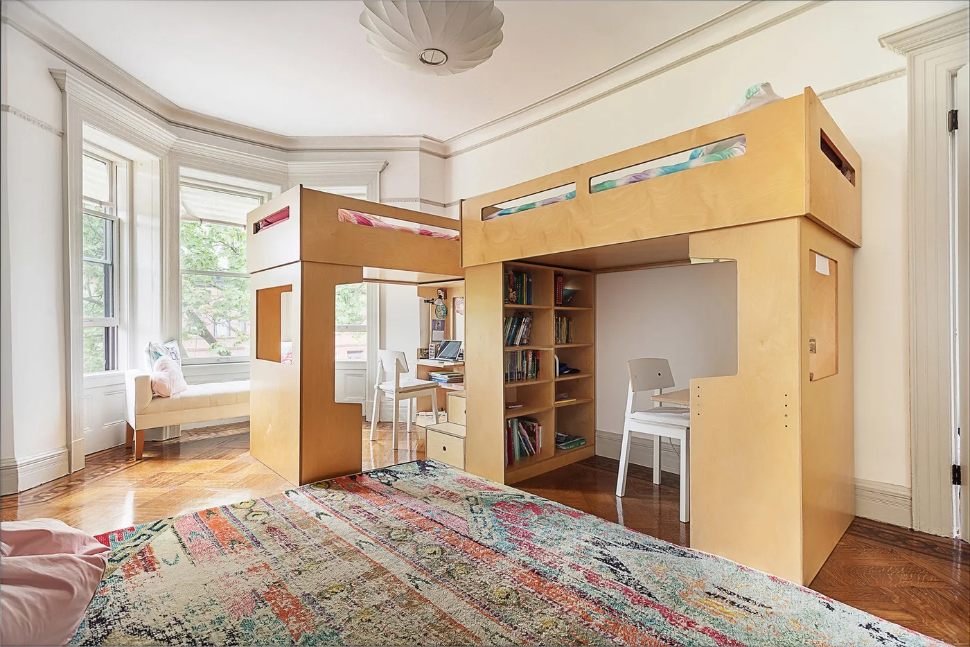 Two pale wood loft beds in an L-shaped arrangement with separate desks, white chairs and a shared central stair and shelving section.