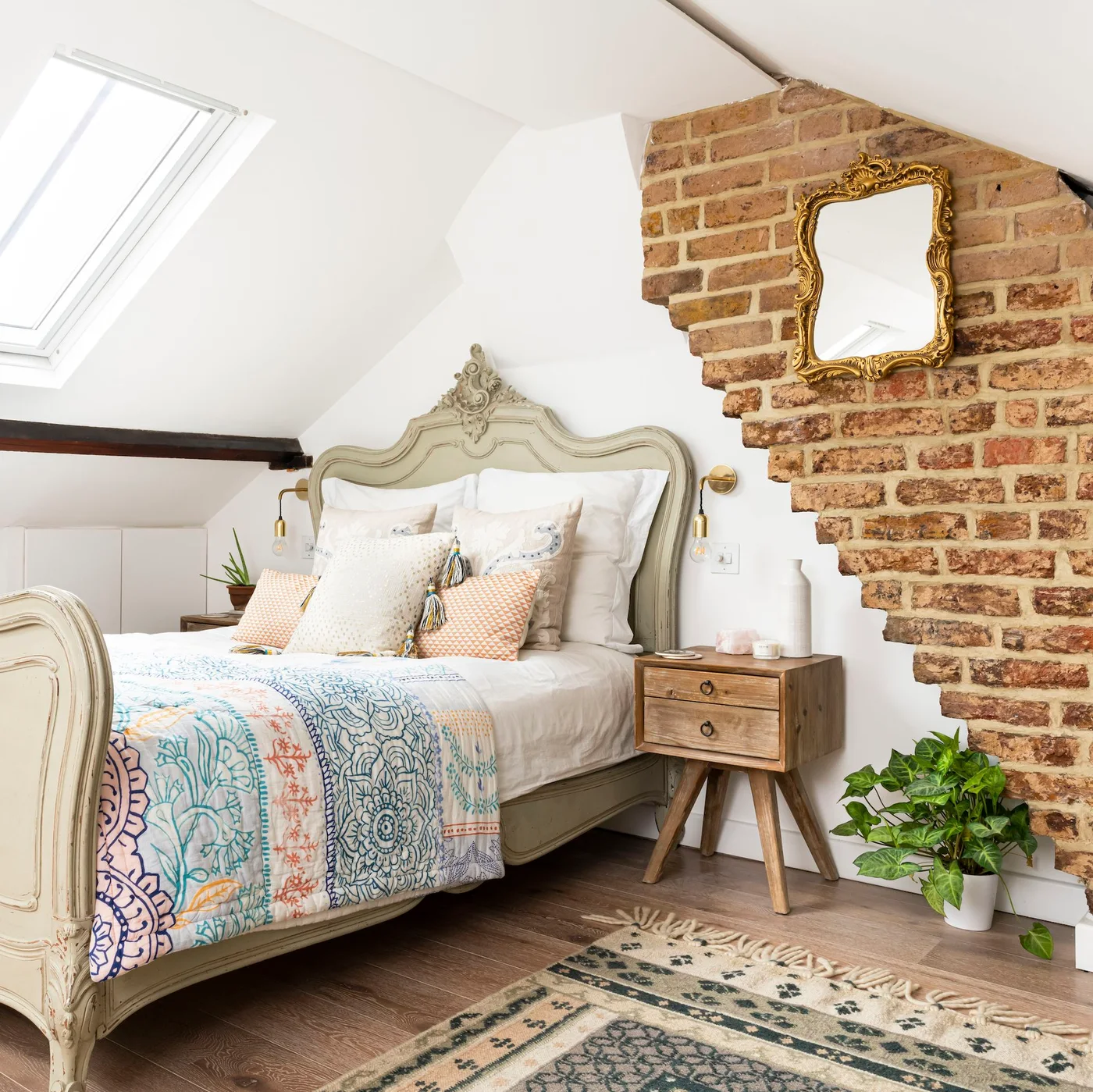 Exposed brick section beside an ornate pale bed in a white attic bedroom with colorful bedding.