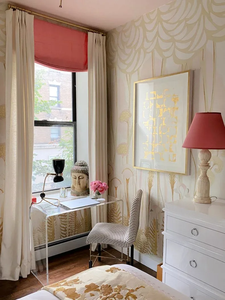 A transparent desk and striped chair stand beside a bedroom window with patterned wallpaper, a pink blind and pale curtains.