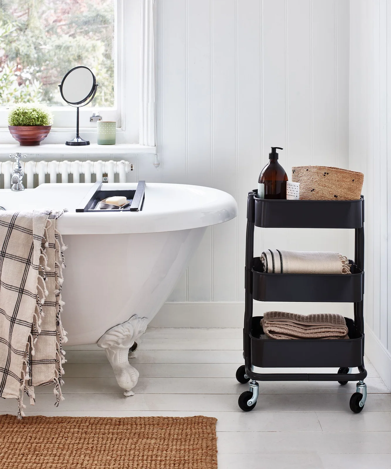 Slim black three-tier trolley with toiletries and folded towels beside a bath