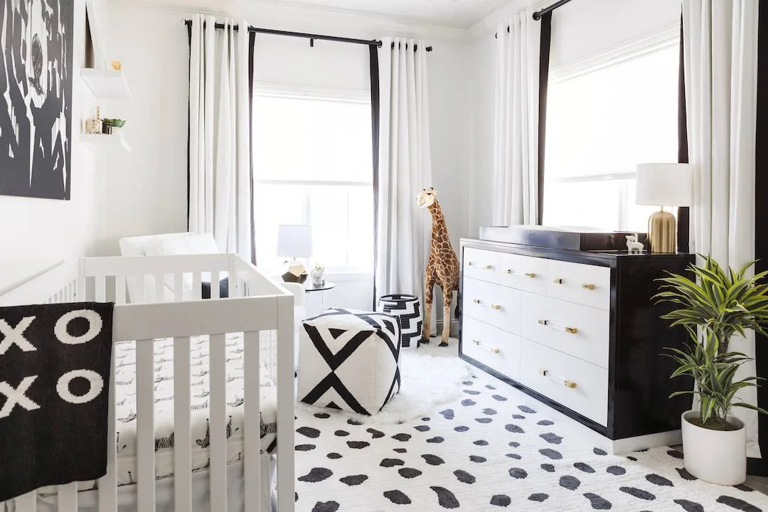 A white nursery crib and chair sit beside a white drawer chest with dark edges, black-and-white spotted rug and light curtains with dark borders