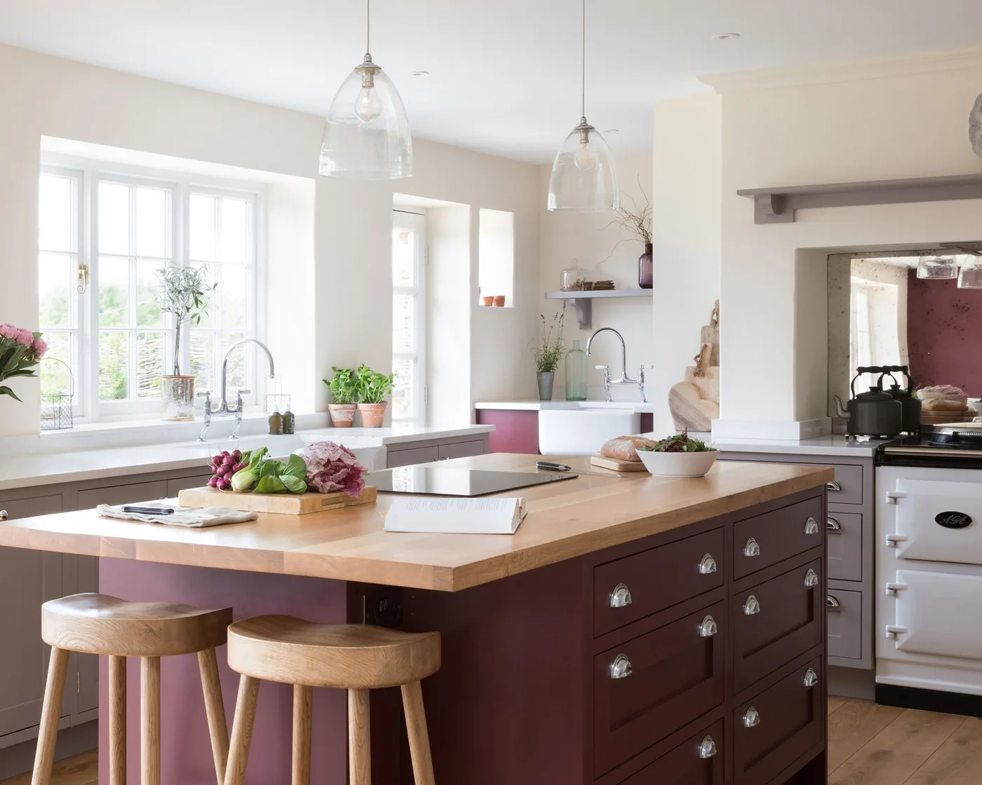 Pale kitchen with a burgundy drawer island, wooden worktop, round wooden stools and two clear pendant lights.