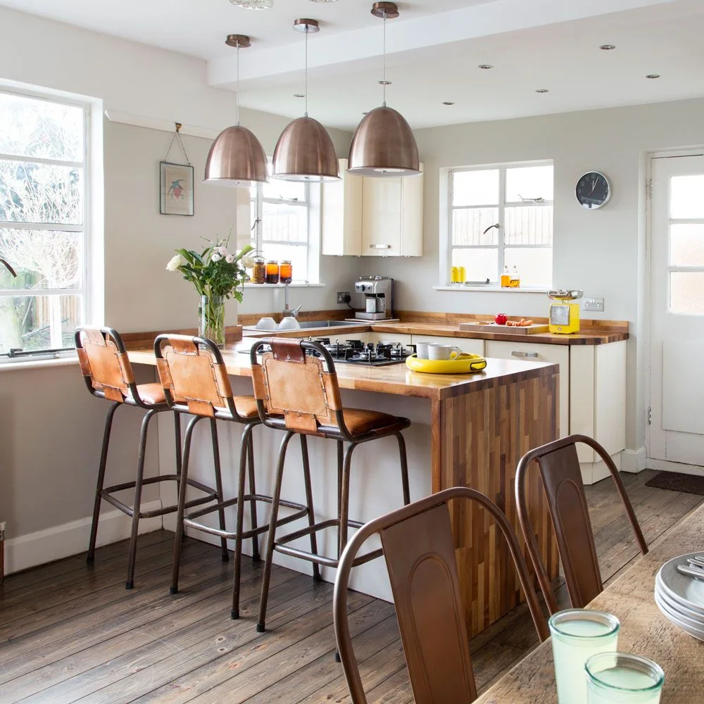 A wooden-topped kitchen island with three brown stools stands beside a dining table beneath copper-toned pendants