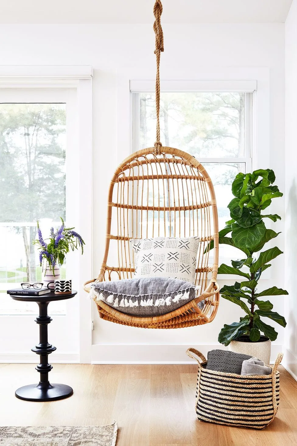 A woven hanging chair with pale cushions sits beside a leafy floor plant, small dark round table and basket of blankets near two bright windows