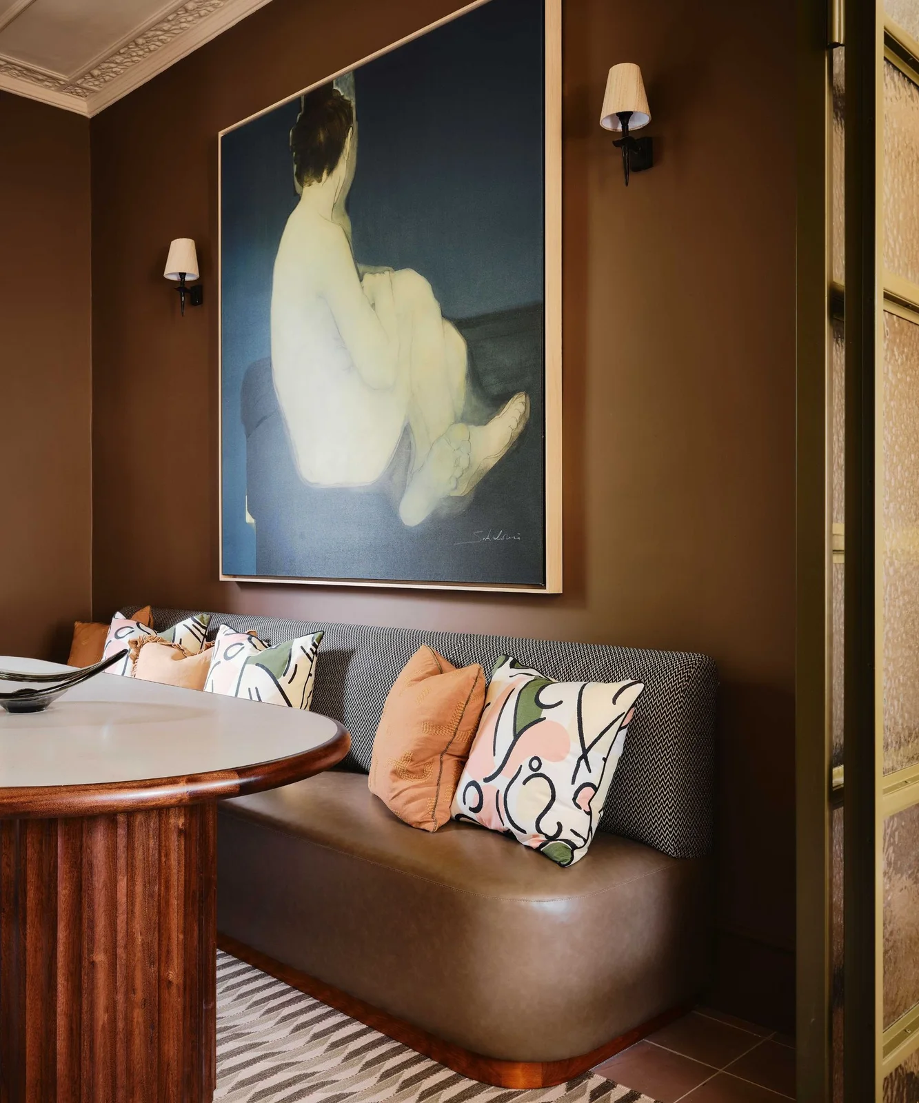 A curved upholstered dining bench sits beneath large artwork and wall lights beside a rounded pale table with a fluted wooden base