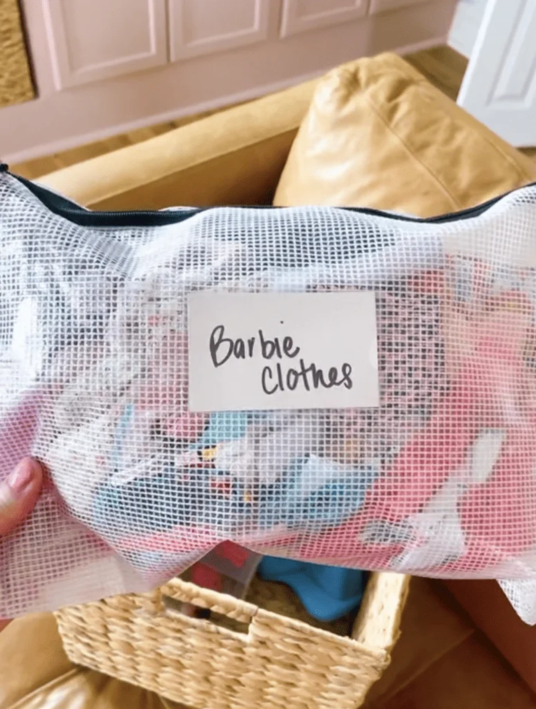 White mesh zipped pouch labeled Barbie Clothes with colorful doll clothing visible inside.