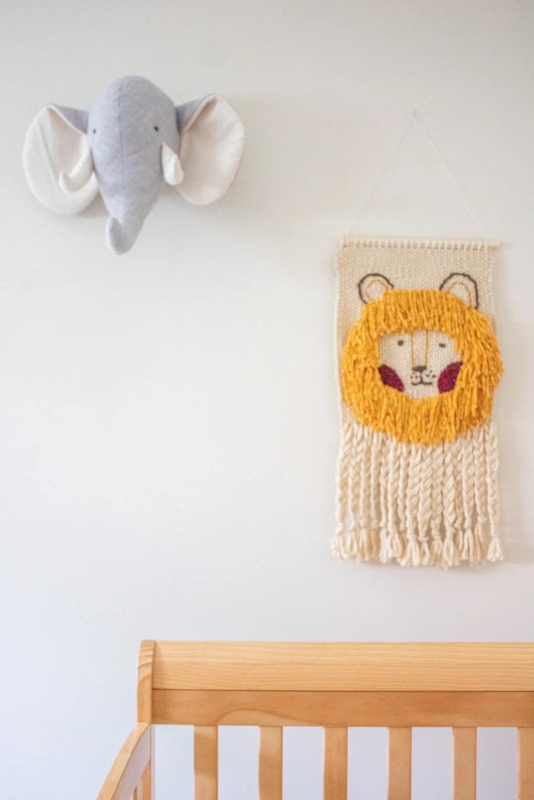 A soft elephant-head wall decoration and a fringed lion tapestry hang against a pale wall above a timber crib rail.