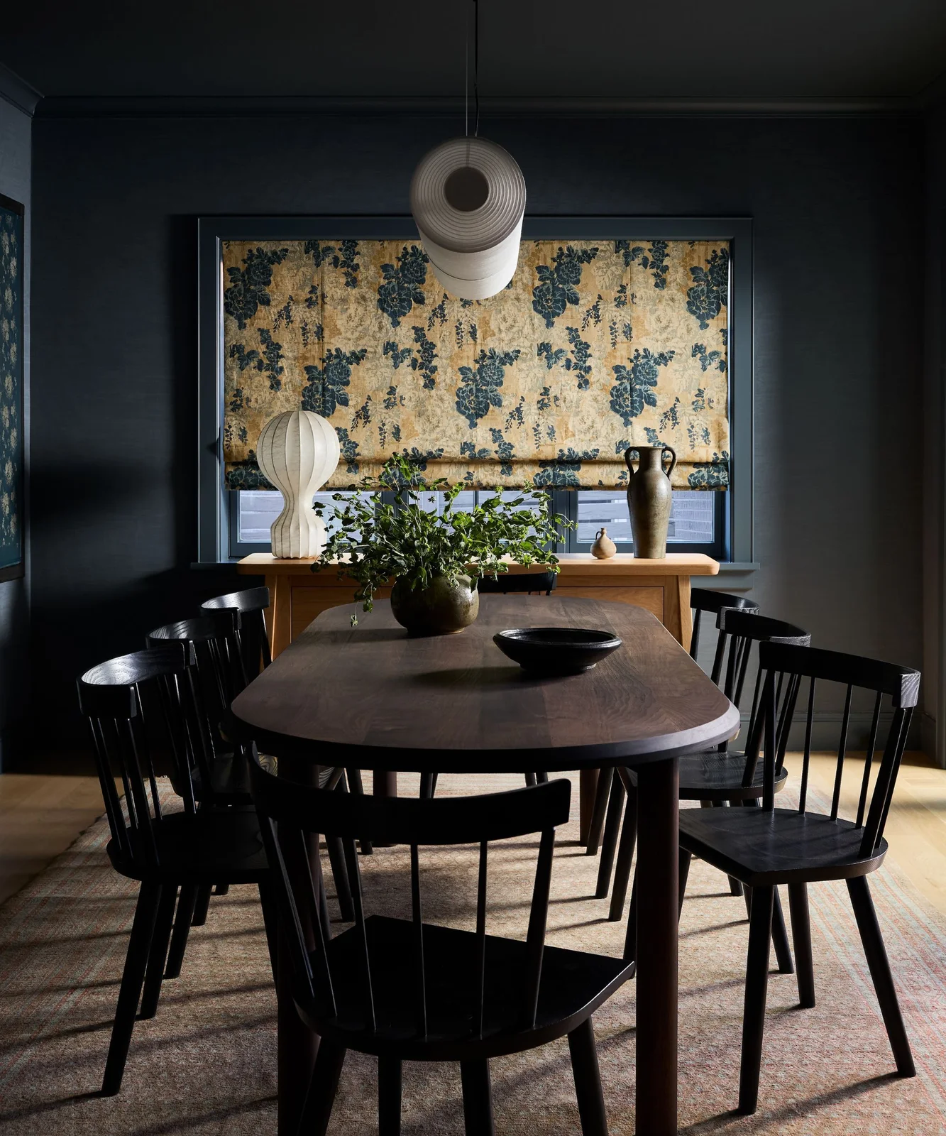 Deep blue walls and ceiling surround a wooden dining table, dark chairs and floral window blind beneath a sculptural pale hanging light
