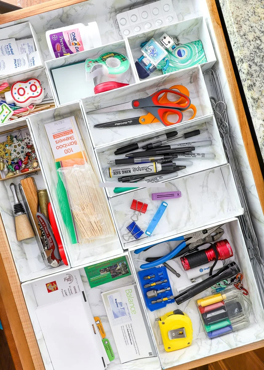 Open drawer divided by marble-pattern boxes containing scissors, markers, tape and household tools