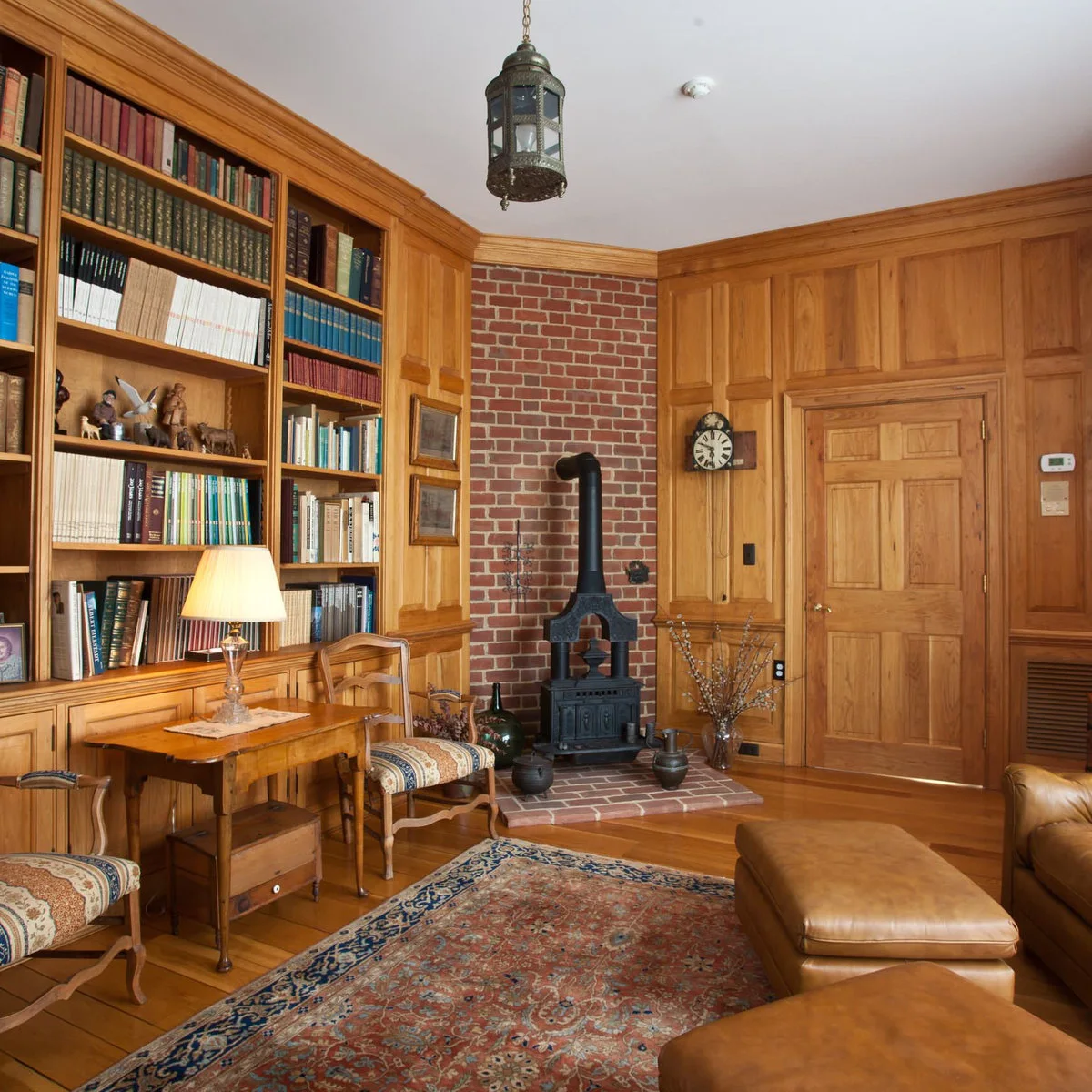 A small dark stove stands before a tall red brick panel in a timber-paneled library with bookshelves, a desk and patterned rug.