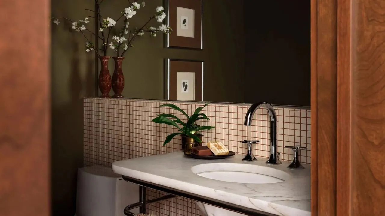 Small square-tile backsplash beneath a dark faucet with framed art and a plant beside a pale basin