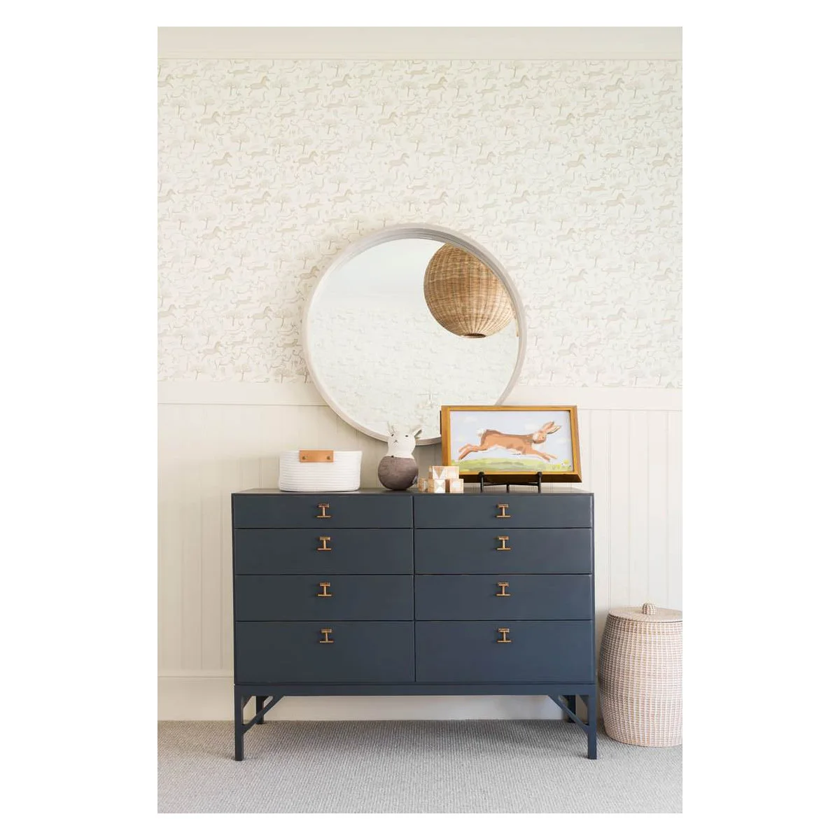 Dark blue nursery dresser below a round mirror, pale animal wallpaper and white paneling