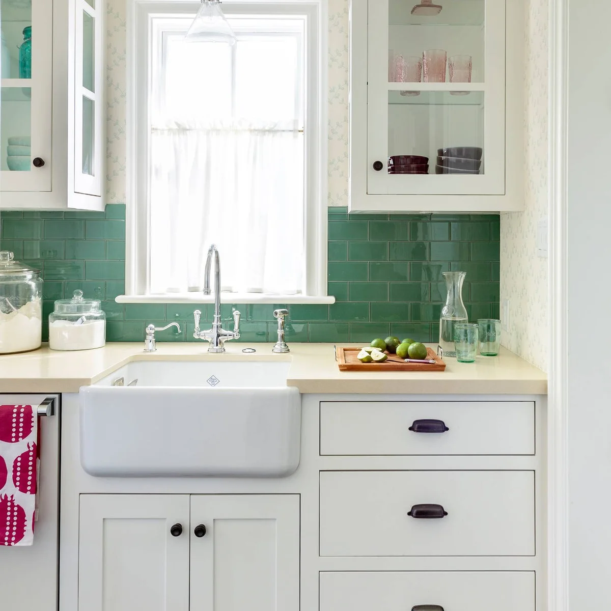 White café curtain above a farmhouse sink between white cabinets and green tiles