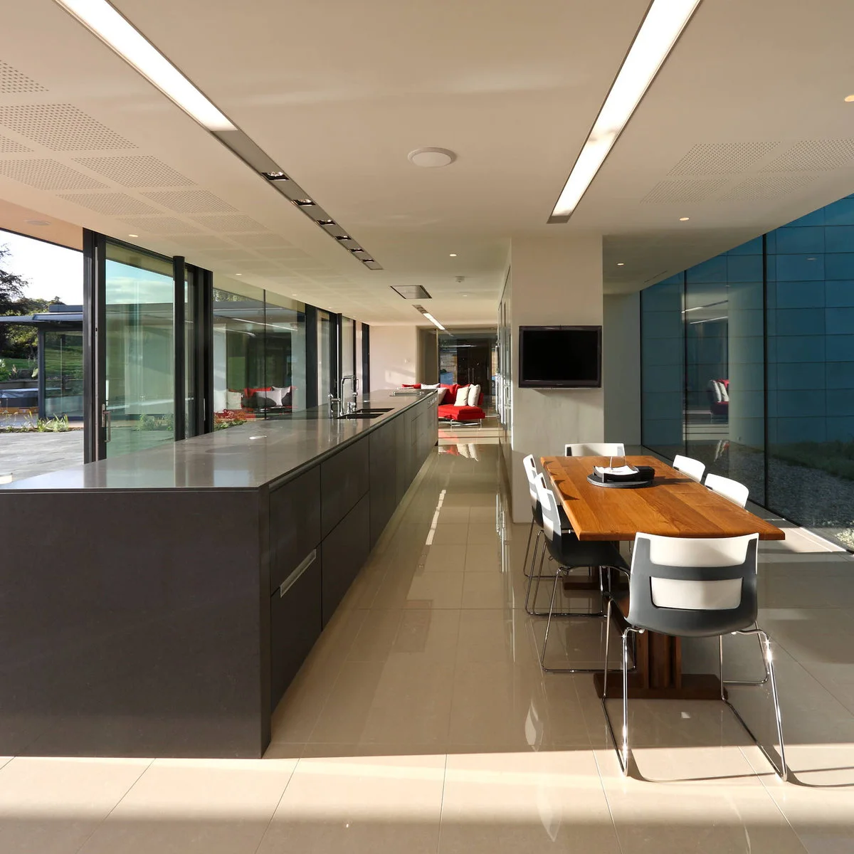 Long glazed room with a dark kitchen island on the left, a wooden dining table on the right and red lounge seating at the far end.