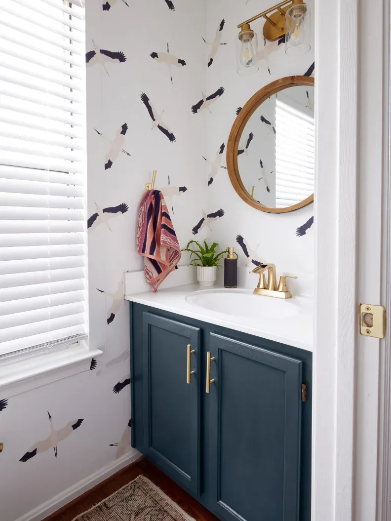 Powder room basin area with white bird-patterned wallpaper, blue vanity, round wooden-framed mirror, warm metallic fittings and striped pink hand towel.