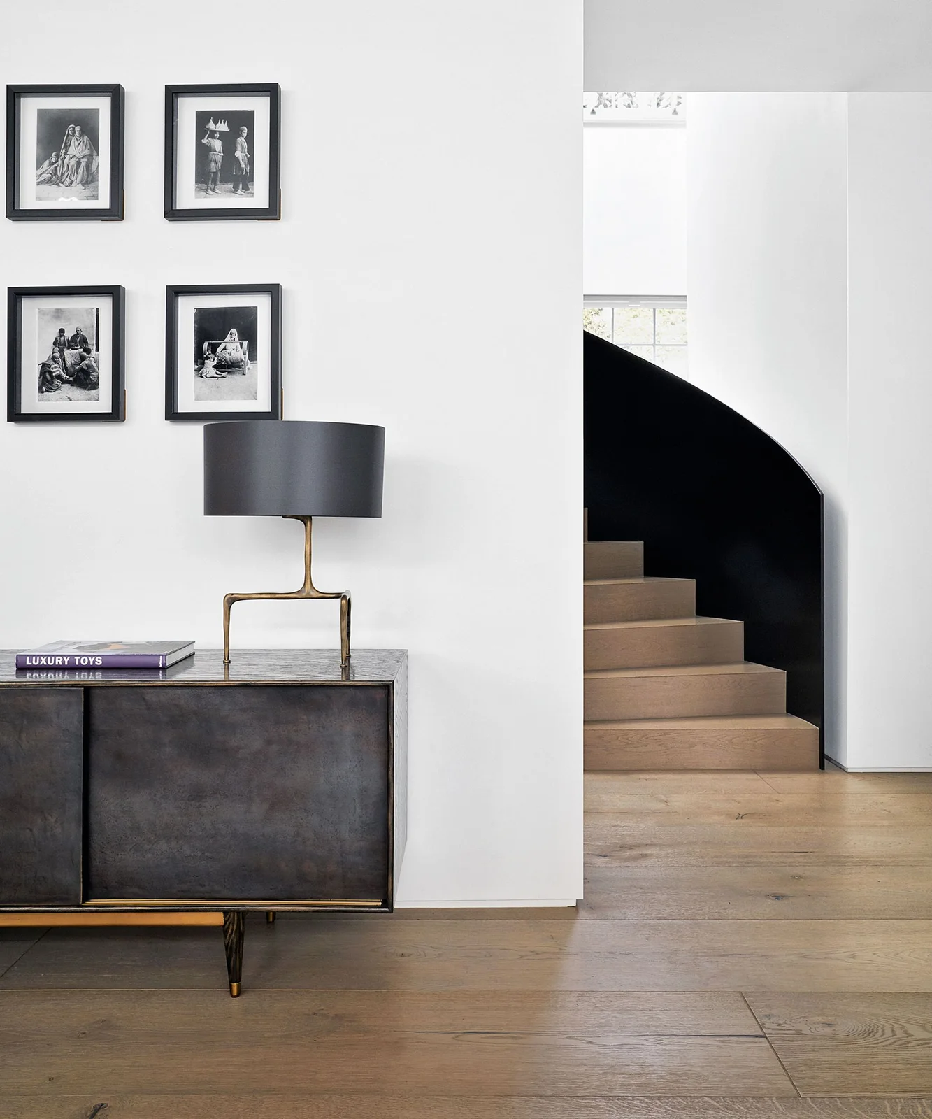 A dark gray cabinet and shaded lamp stand beneath four black-framed pictures beside white walls, a curved black stair enclosure and warm wood flooring