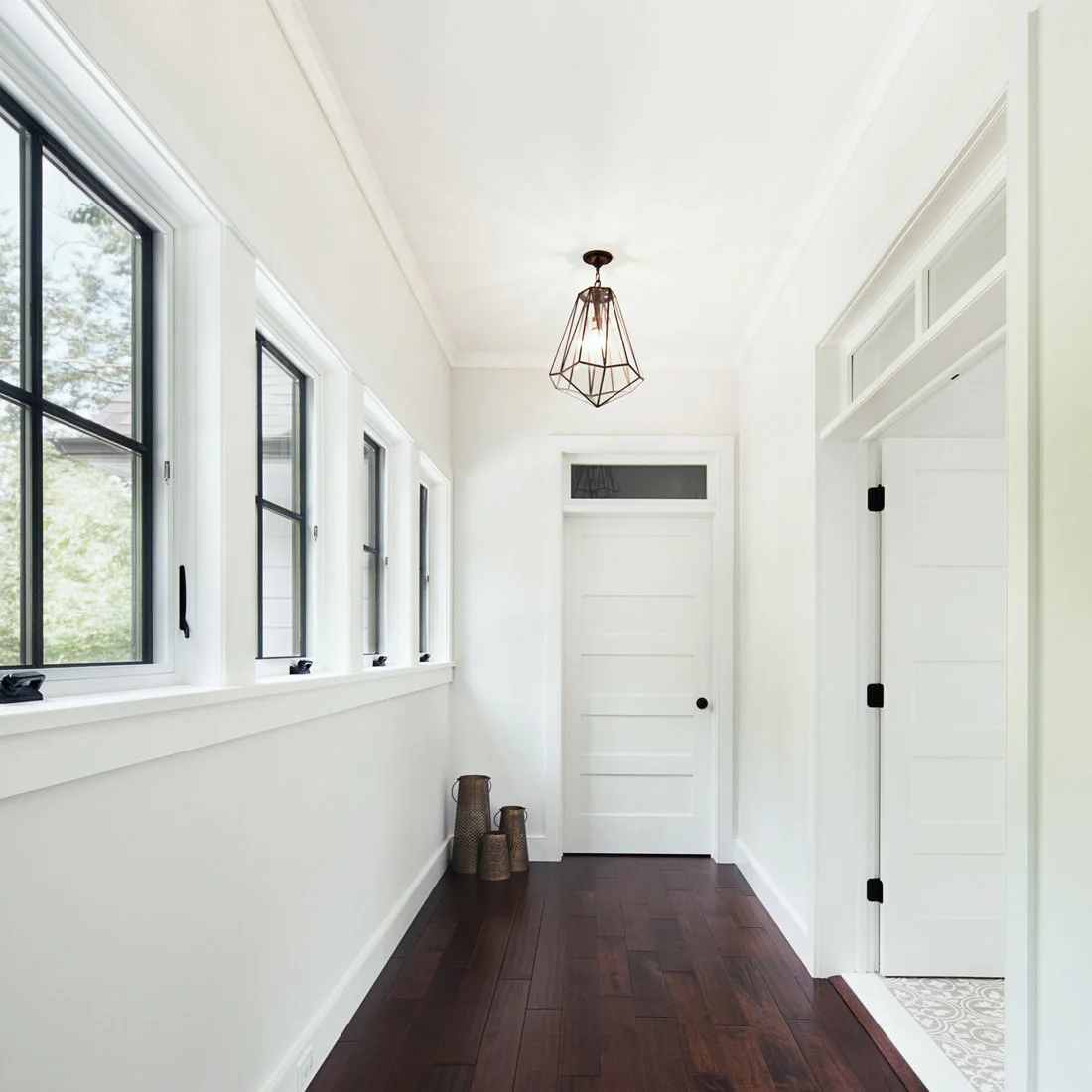 A white hallway with black-framed windows, white doors and transoms has a dark wood floor and a geometric pendant.