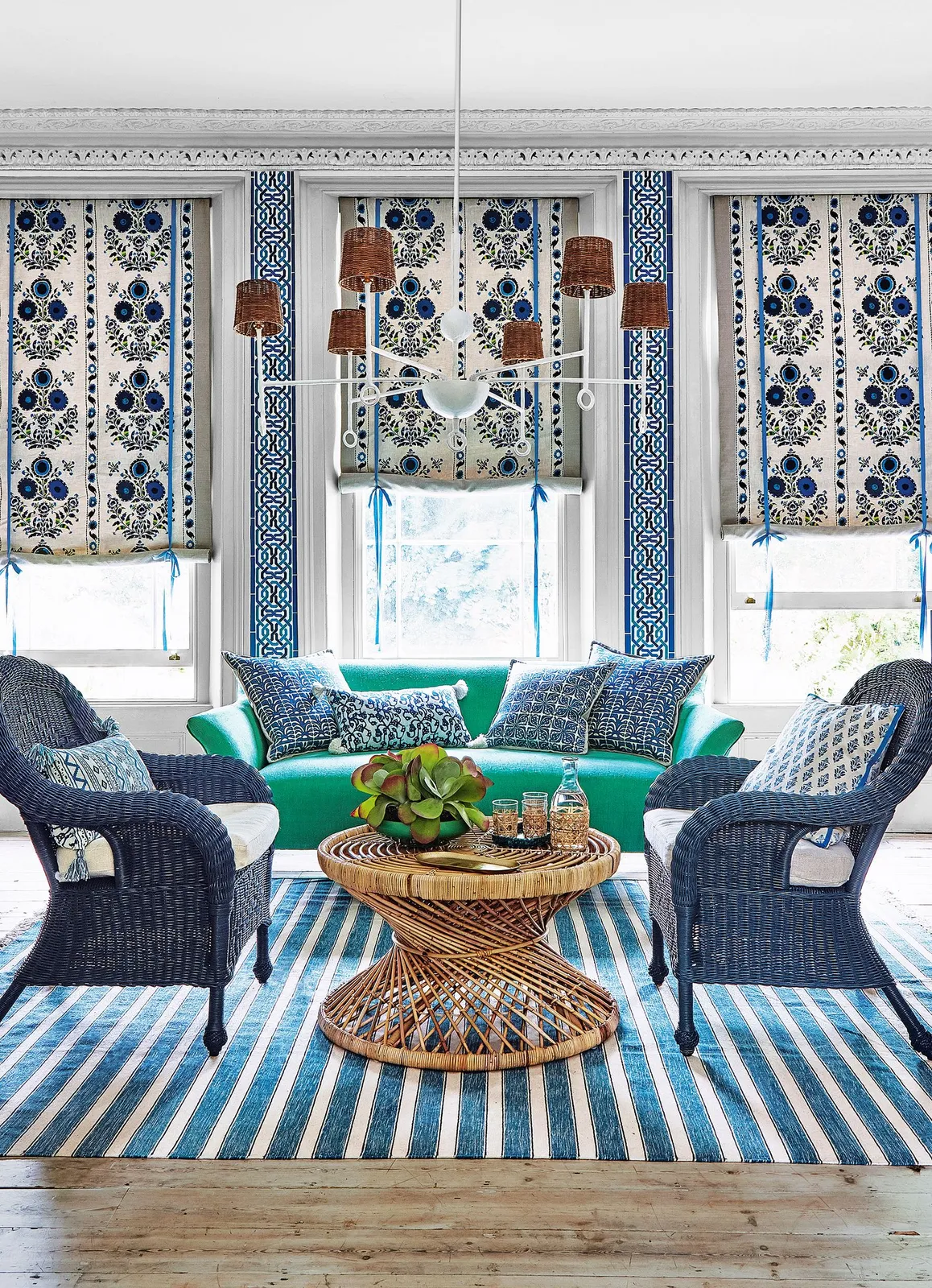 Living room with blue-and-white vertical wall patterns and blinds, blue woven chairs, green sofa, striped blue rug and round woven table.