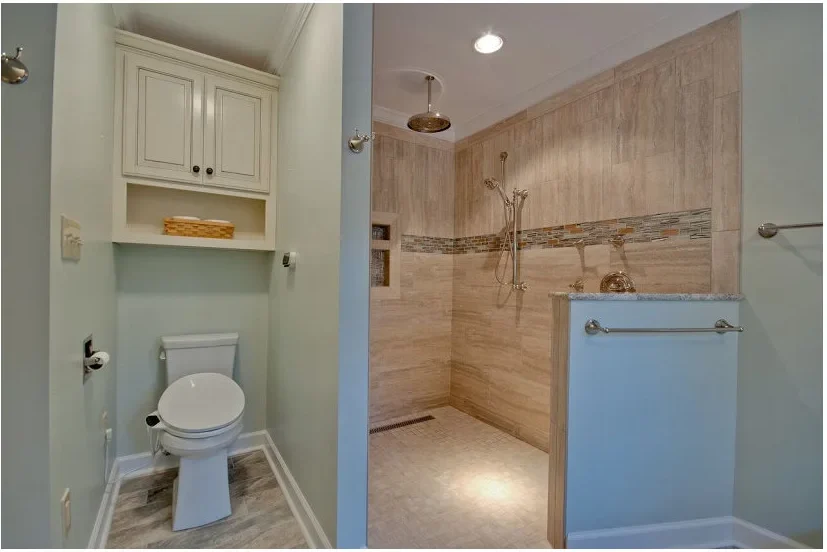 A pale blue half wall borders an open shower entrance beside a toilet alcove, with beige tiles and a ceiling-mounted showerhead.