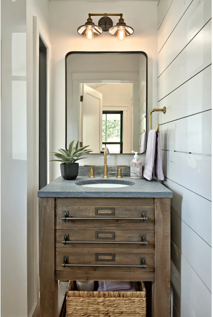 A brass-colored angular towel holder sits on a white paneled side wall beside a narrow wood vanity and dark counter.