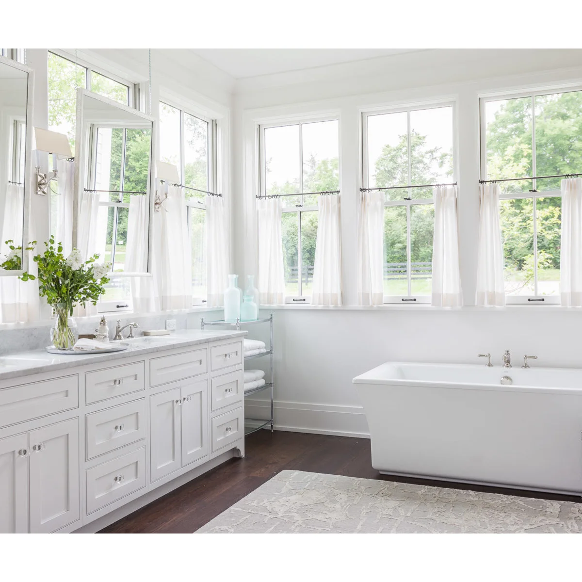 White cafe curtains repeat across tall windows wrapping a bathroom corner above a vanity and freestanding bath.