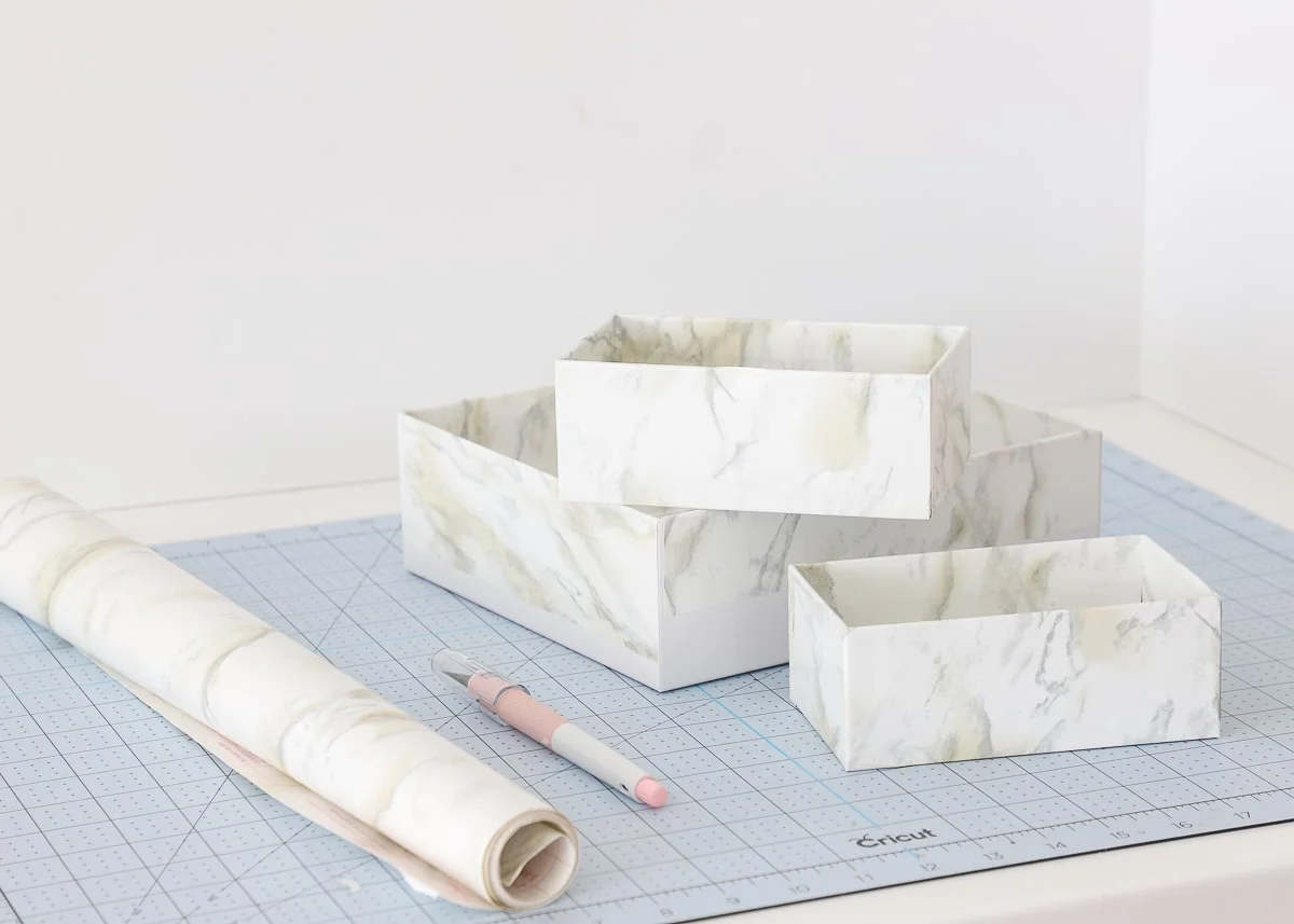 Three marble-pattern covered boxes on a cutting mat beside a roll of covering paper