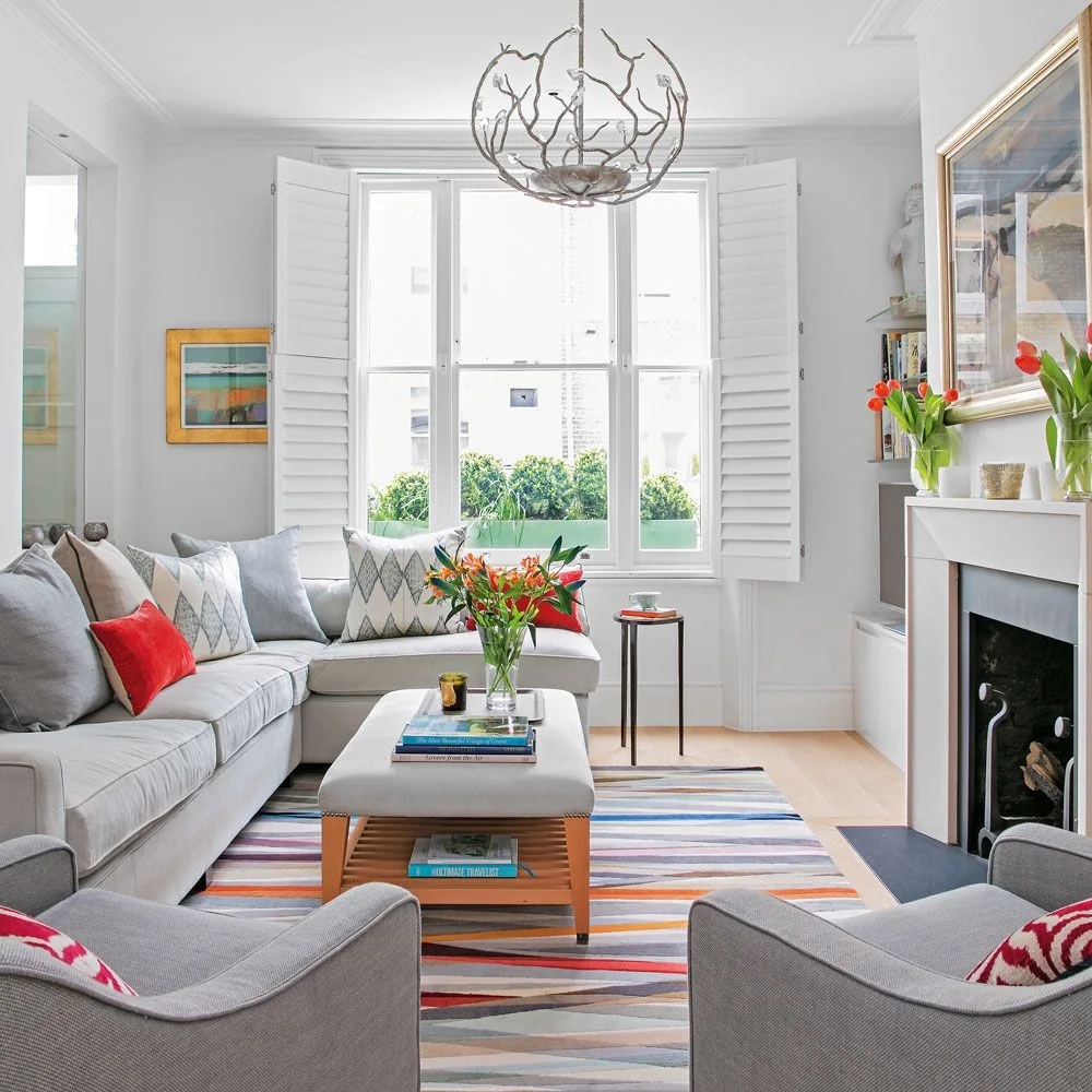 A pale gray sectional, two gray chairs and an upholstered ottoman surround a striped rug near white window shutters and a fireplace