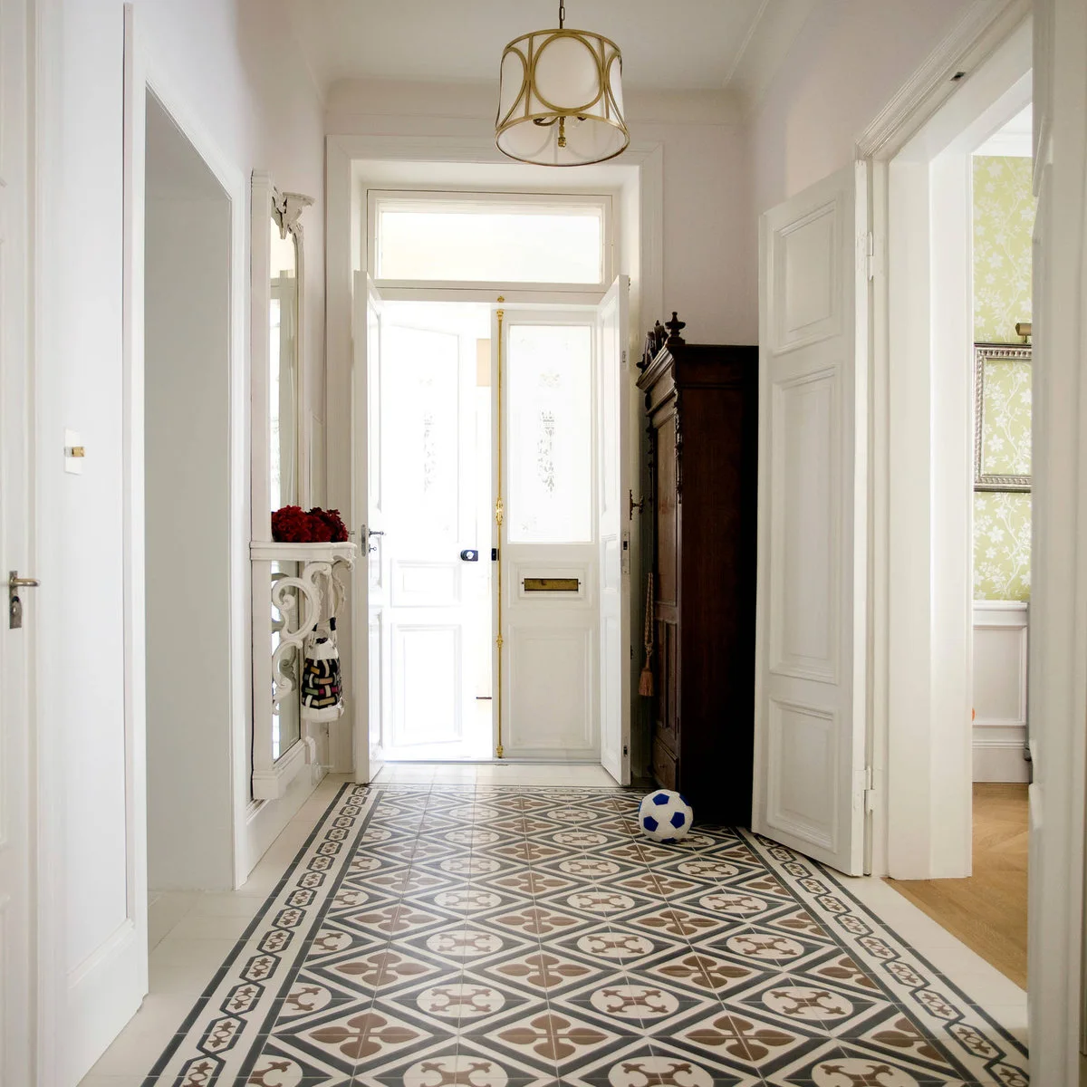 Pale entrance hall with a geometric patterned floor, white glazed front door and tall dark wooden cabinet.