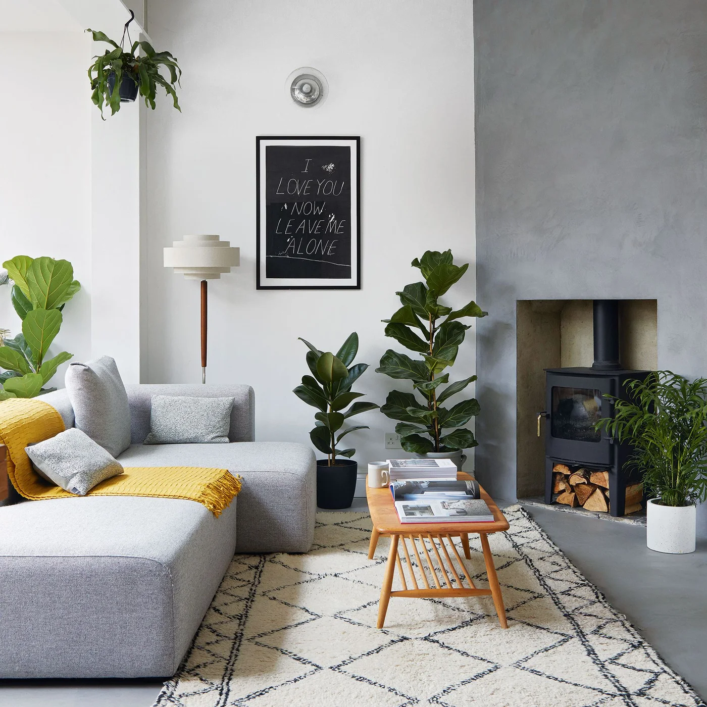 A gray textured chimney wall frames a black stove beside a pale gray corner sofa, wooden tables and leafy plants