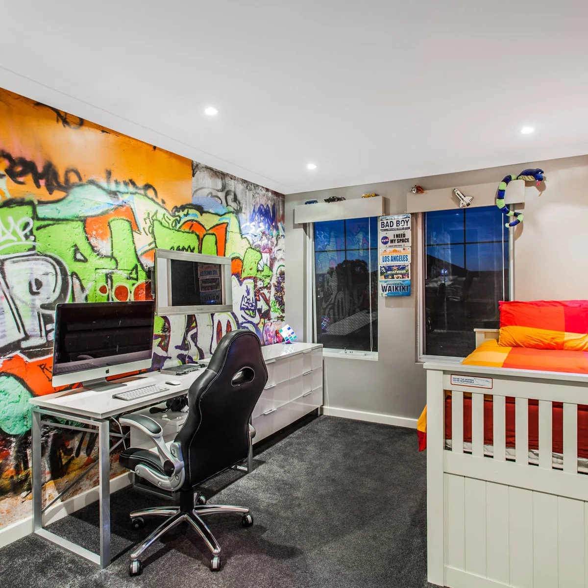 A bedroom with colorful graffiti-style wall art, a black gaming chair, a desk computer and a separate wall television.